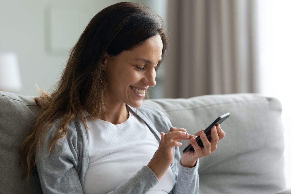 Happy mixed race millennial girl sitting on comfortable coach in living room, using mobile phone applications, shopping, playing online games, chatting with friends, watching funny videos, head shot; property management marketing